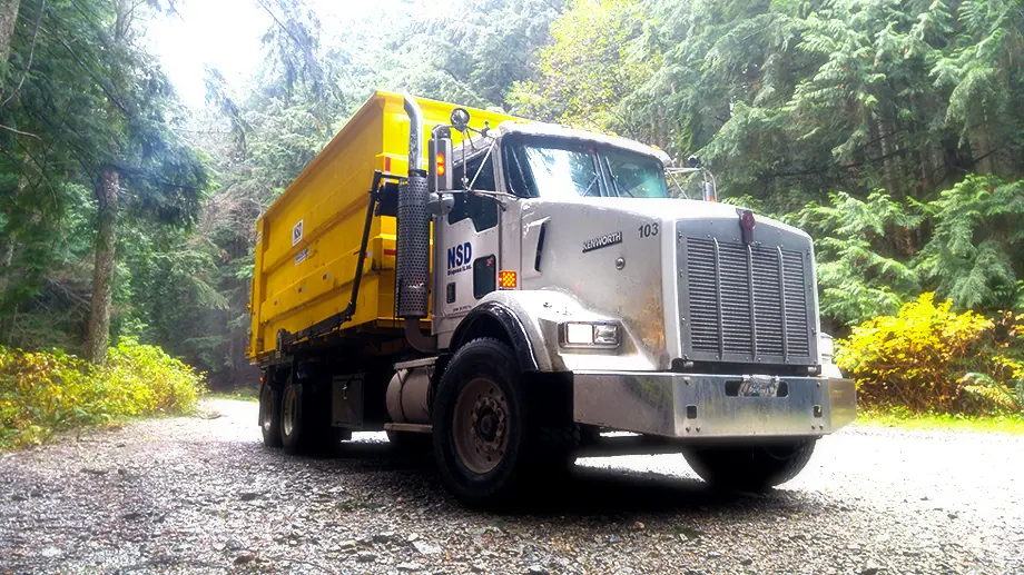 NSD roll-off truck with a yellow bin on a tree-lined gravel road